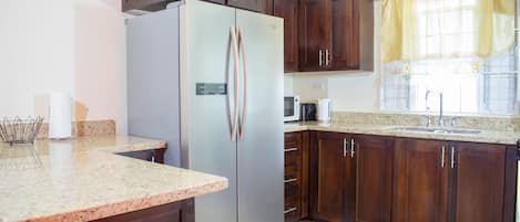 A kitchen with dark wooden cabinets, a stainless steel refrigerator, a granite countertop, and a window with a yellow curtain.