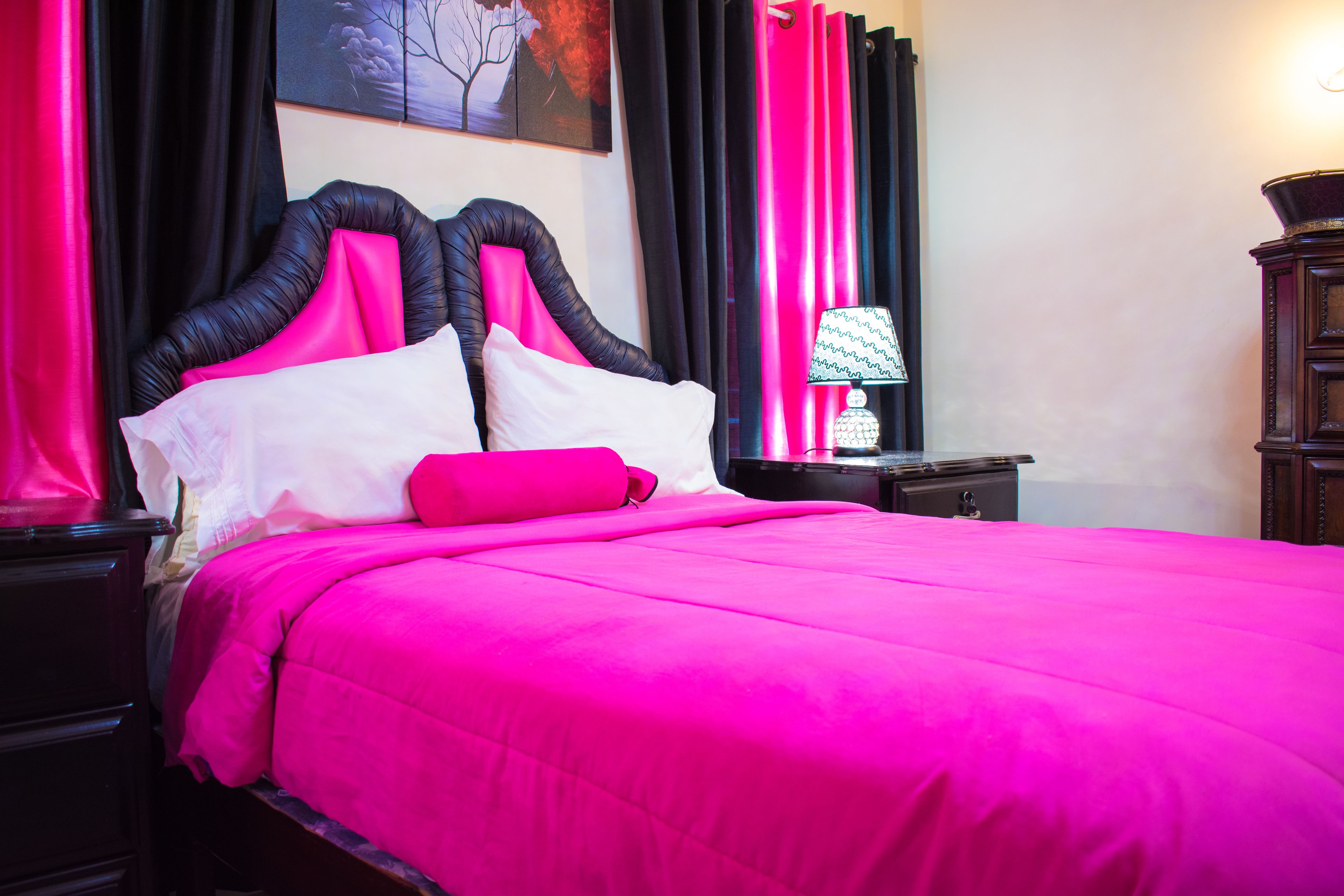 A bed with a black headboard and bright pink bedding, a nightstand with a lamp, and dark curtains.