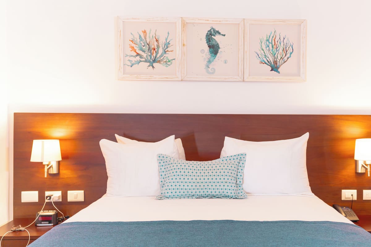 Une chambre d’hôtel avec un grand lit, deux lampes de chevet, une table de chevet avec un téléphone et trois tableaux représentant des hippocampes, encadrés et accrochés au mur.