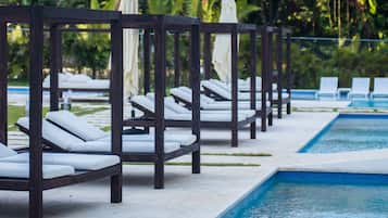 Un área junto a la piscina con sillas de descanso y sombrillas, rodeada de vegetación exuberante y palmeras.