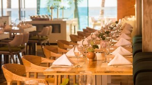 Un comedor con mesas de madera, servilletas blancas y cristalería, con vistas a un paisaje con palmeras y el océano.