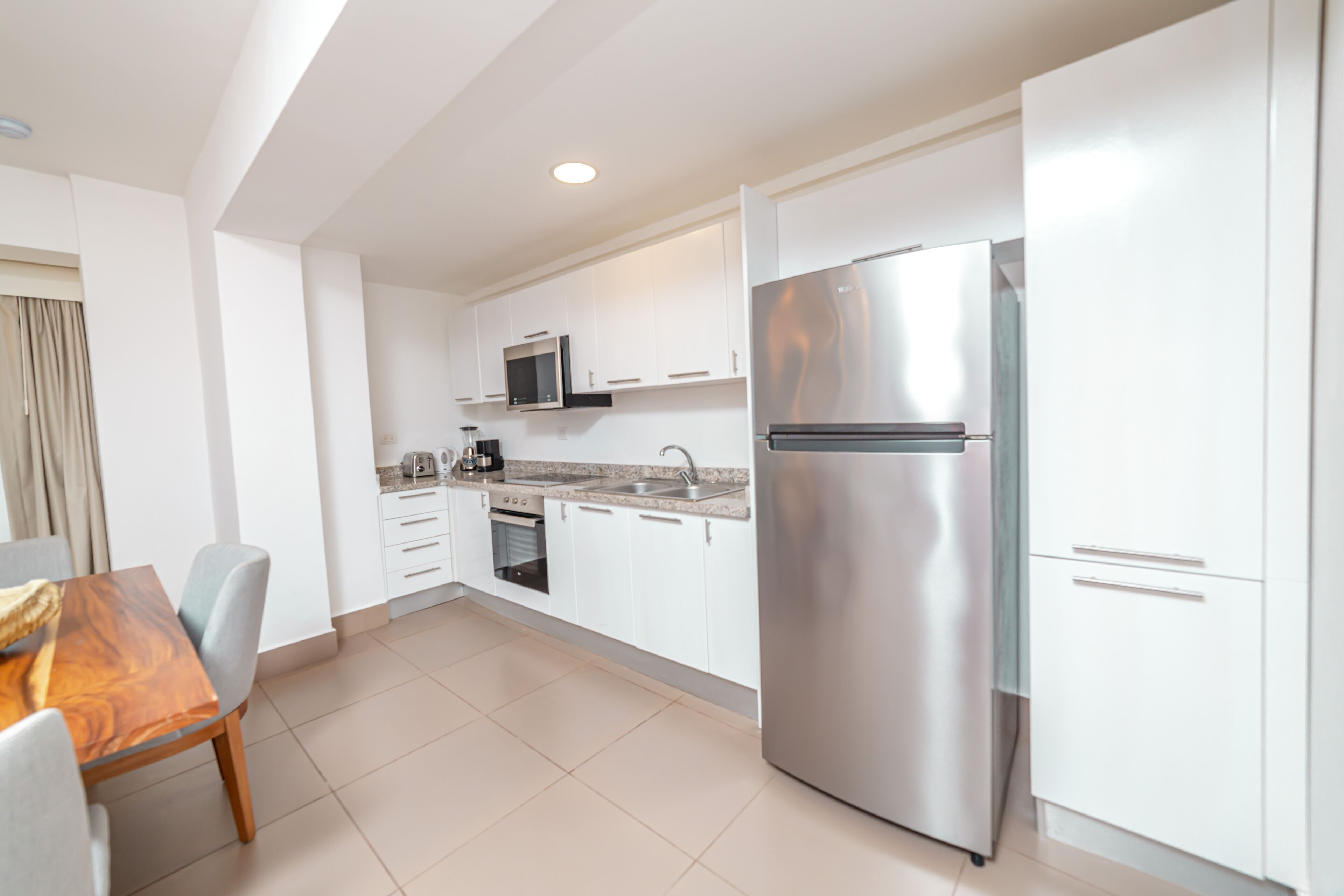 A modern kitchen with white cabinets, a stainless steel refrigerator, a microwave, and a dining area with a wooden table and chairs.