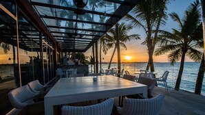 Un área de comedor al aire libre con mesa y sillas, con vista a una playa con palmeras y el océano al atardecer.