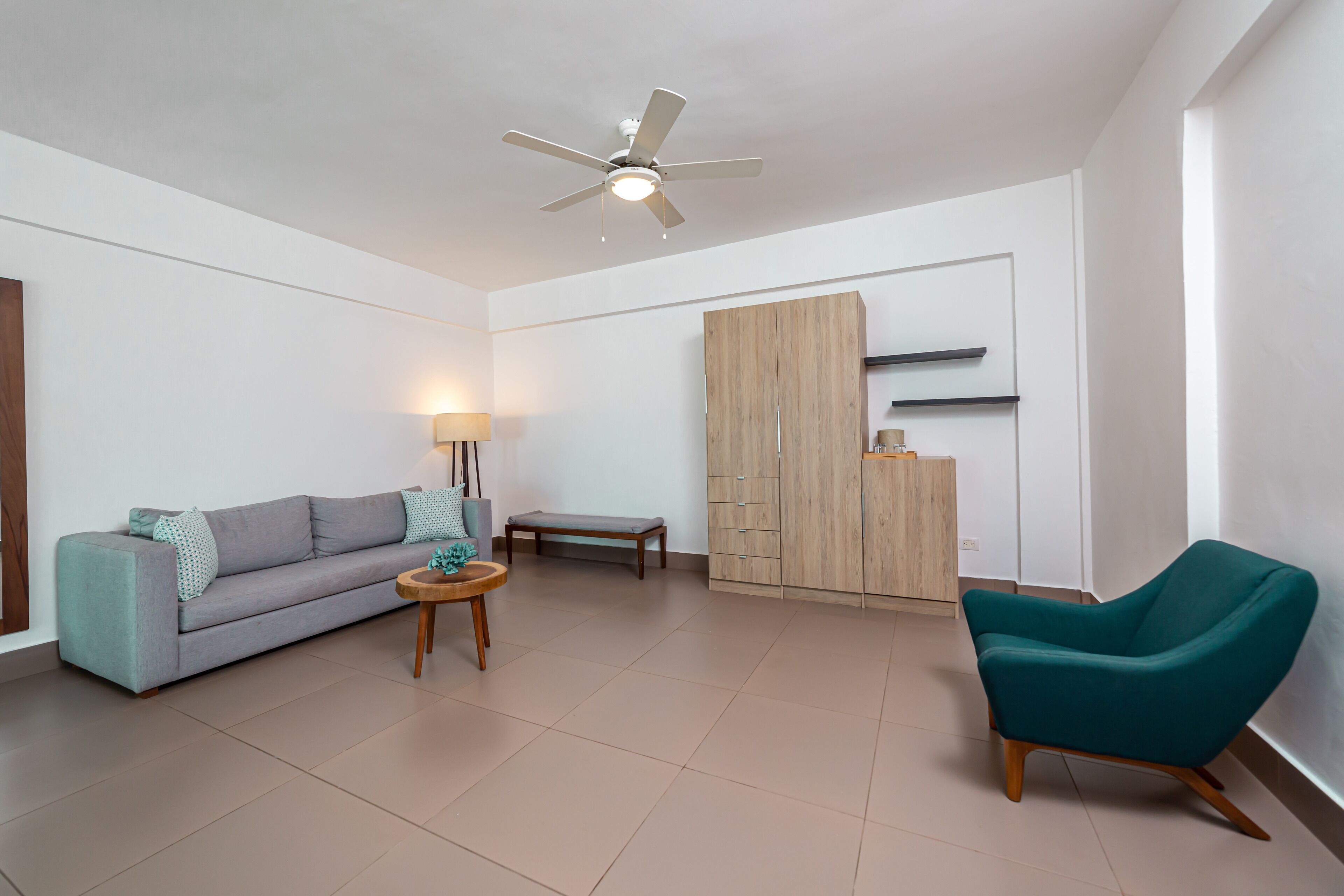 Ein modernes Wohnzimmer mit Sofa, einem petrolfarbenen Sessel, einem kleinen Tisch, einer Stehlampe, einem Deckenventilator und einem Kleiderschrank.