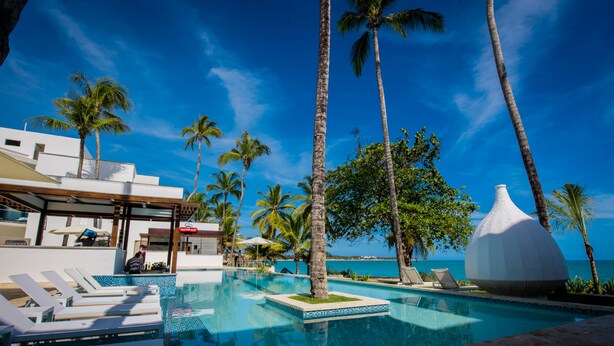 Zona de piscina con sillones de descanso, palmeras y vista al océano.
