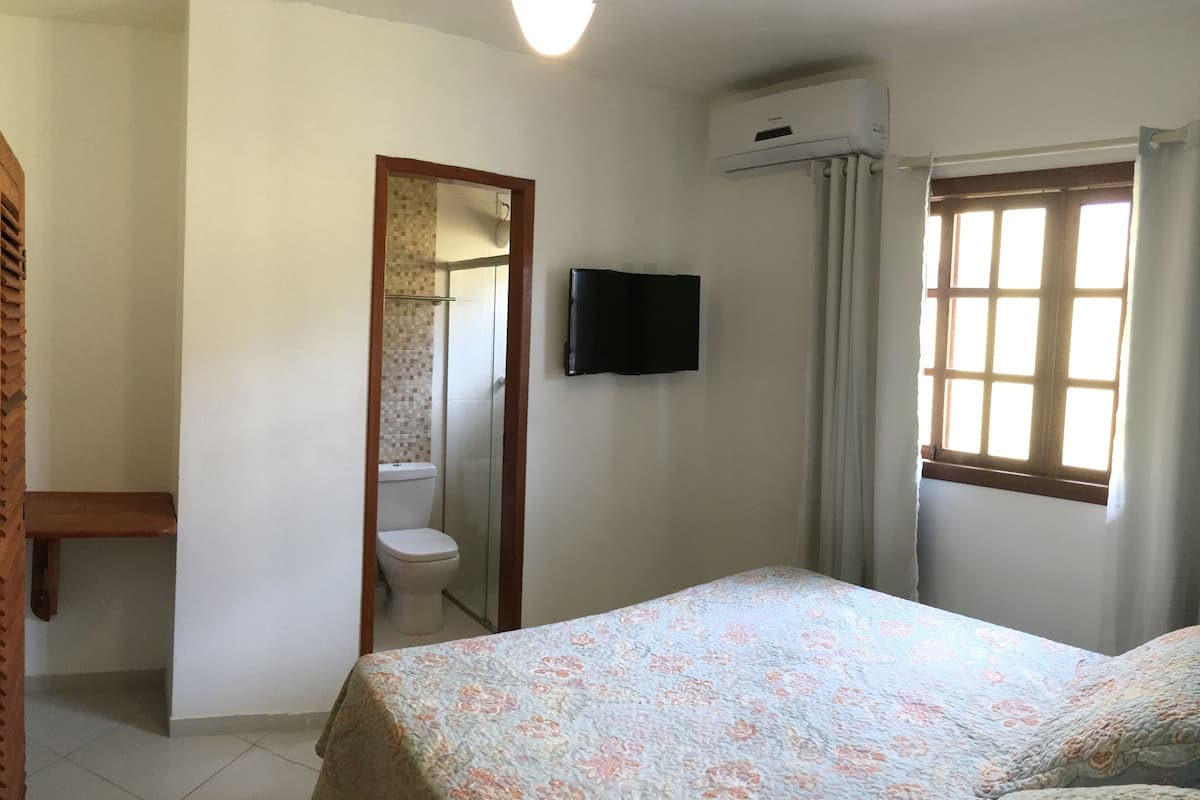 Ein Schlafzimmer mit einem Bett, einem Fernseher, einem Fenster mit Vorhängen und einem durch eine offene Tür einsehbaren Badezimmer.