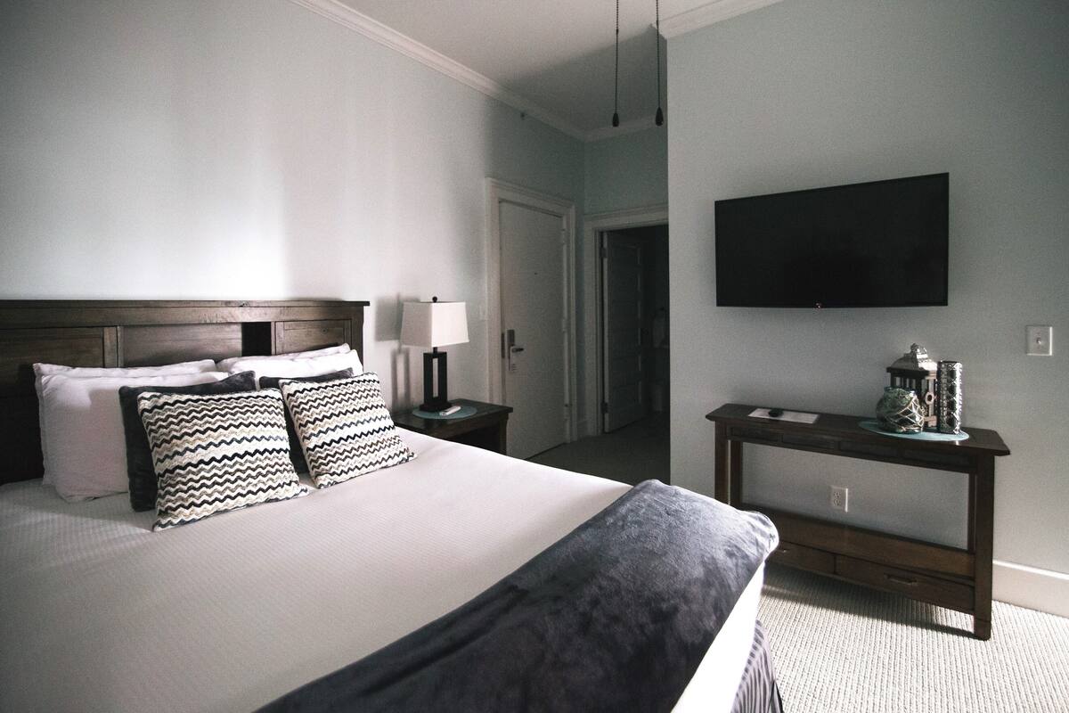 A hotel room with a large bed, a bedside table with a lamp, a TV mounted on the wall, and a console table with decorative items.