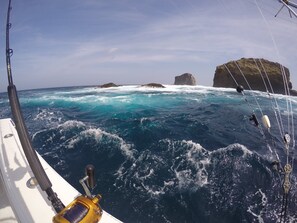 A boat's interior with a fishing rod and tackle, rough sea, and rocky coastline.