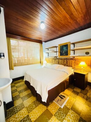A hotel room with a bed, a bedside table with a lamp, a shelf with decorative items, a window with curtains, and a patterned floor. - Hotel Casa Chapultepec - Hostel (Cobán)