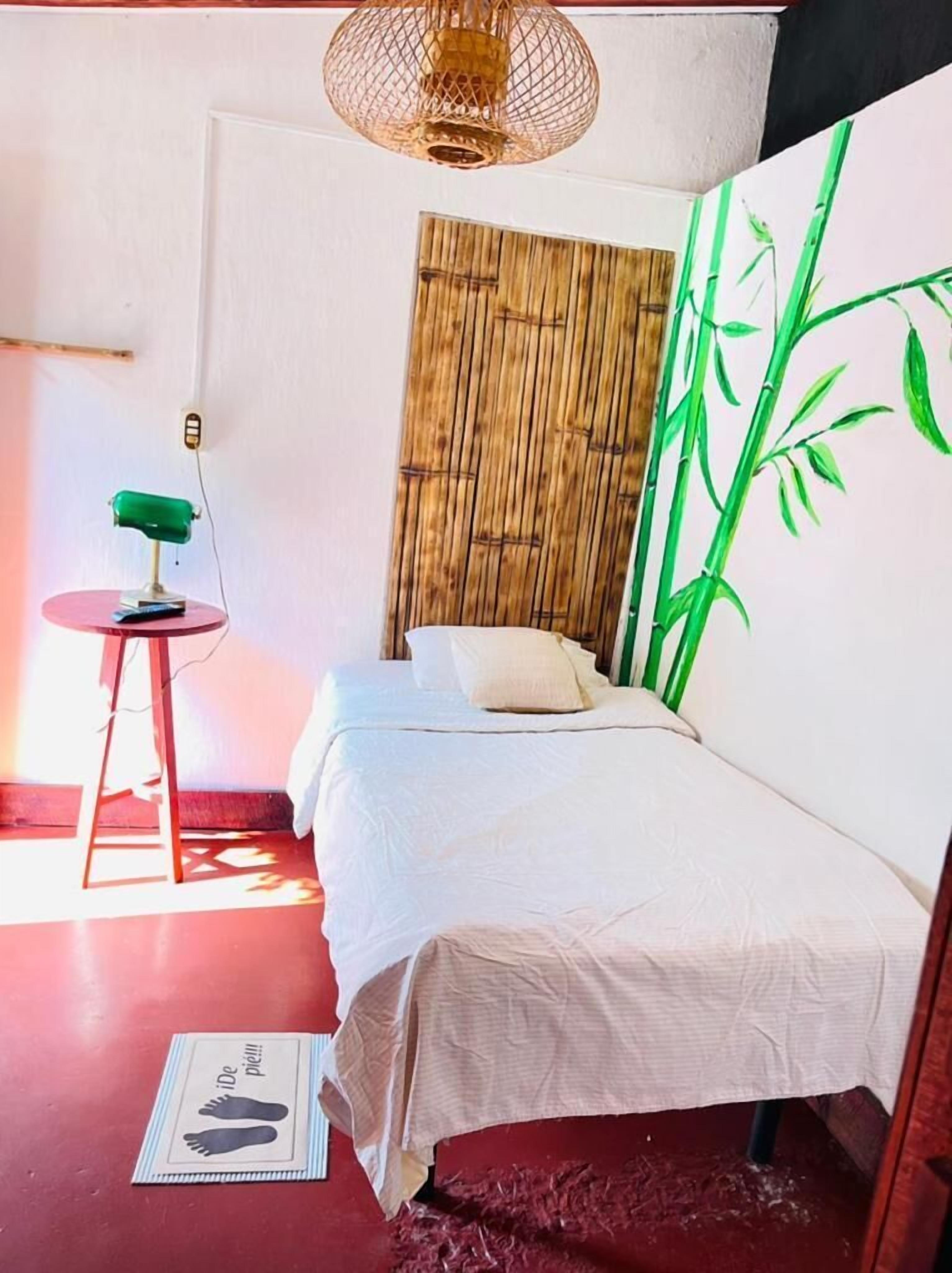 A room with a bed, a small table with a lamp, a wall with a bamboo print, and a hanging light fixture.