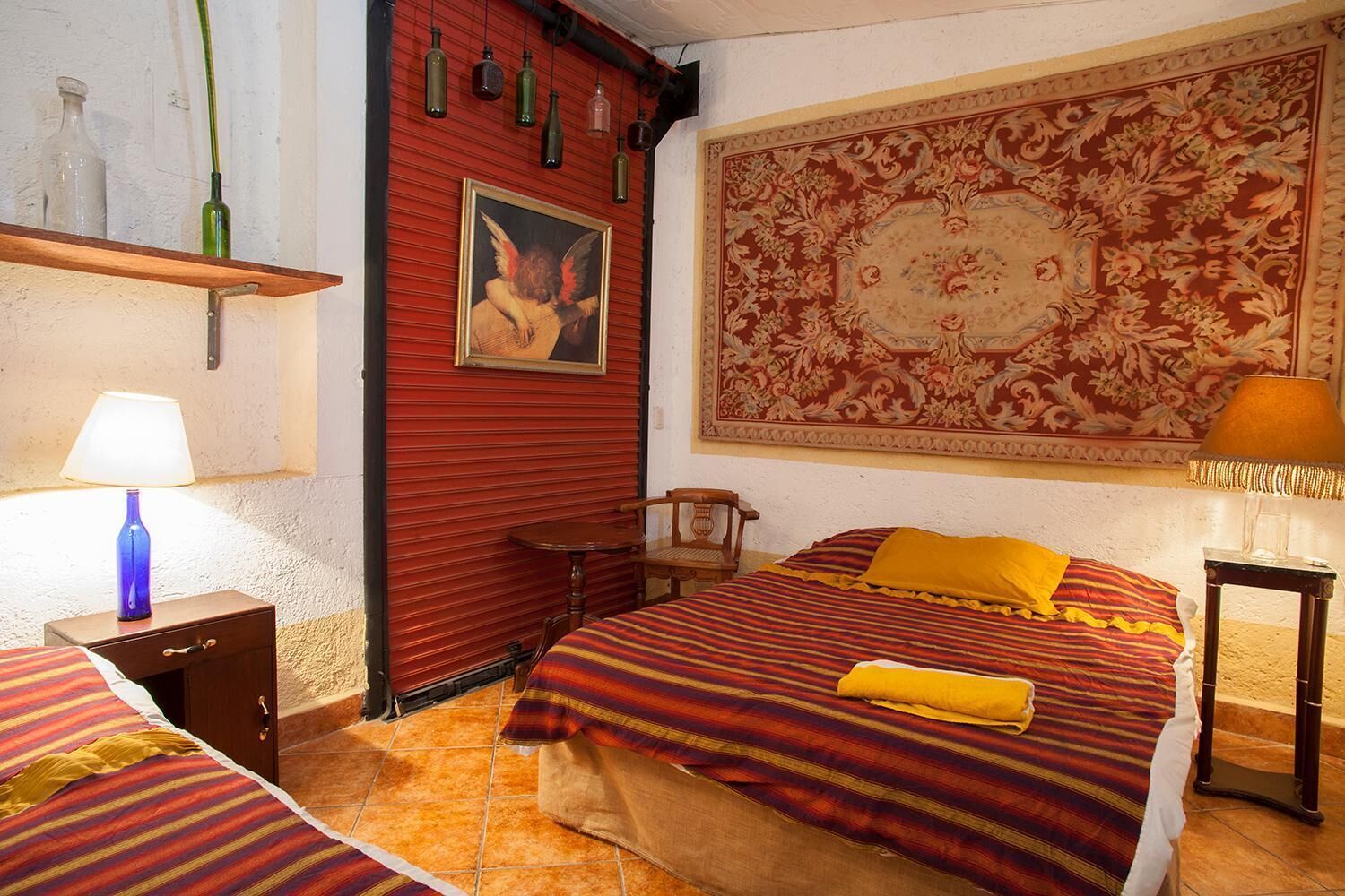 Une chambre d’hôtel avec un mur rouge, une grande tapisserie à motifs floraux, un lit avec une literie à rayures, une chaise en bois et une petite table avec une lampe.