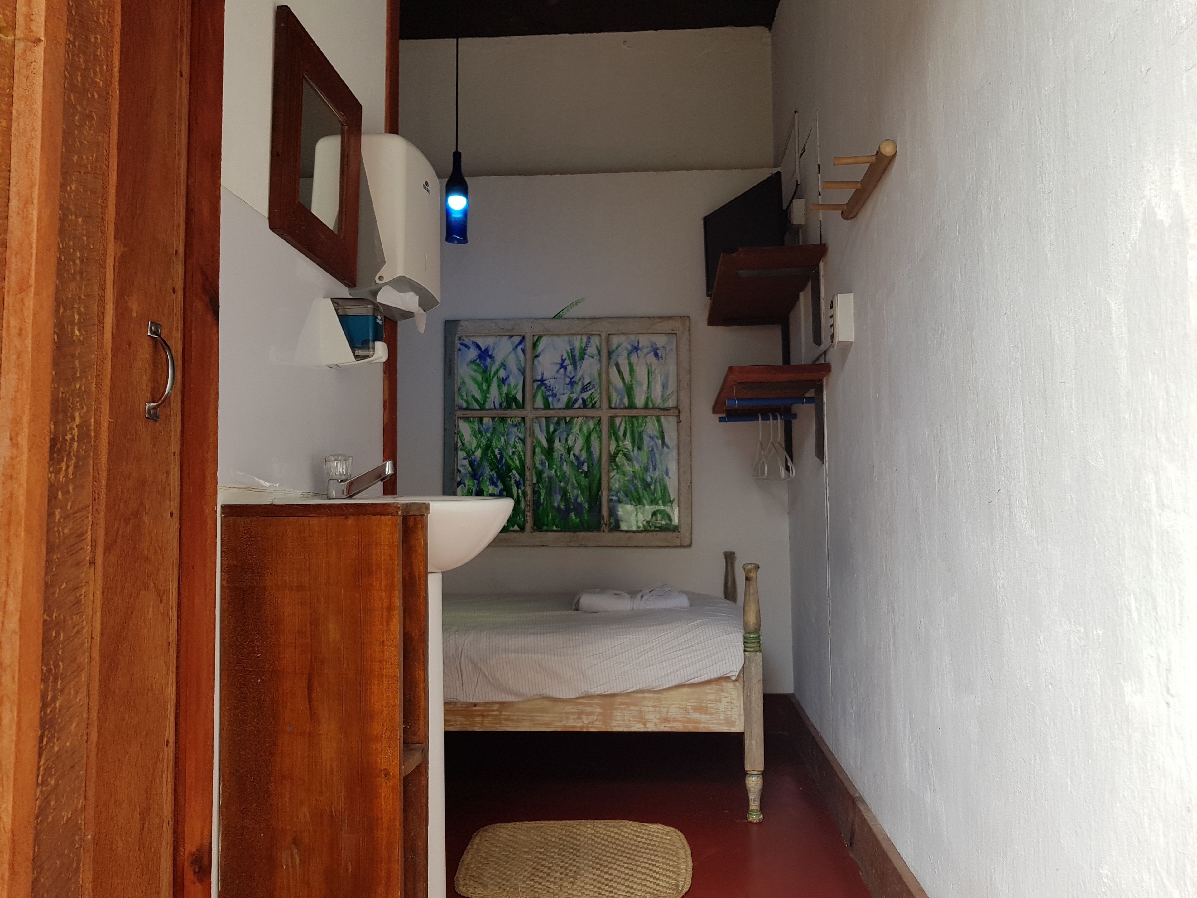 A narrow room with a wooden door, a white sink, a small bedside table, a single bed, a window with a painting, and a hanging light fixture.
