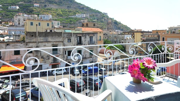 Un balcon avec une balustrade blanche, une table ornée d’un vase de fleurs et une vue sur une colline parsemée de bâtiments.