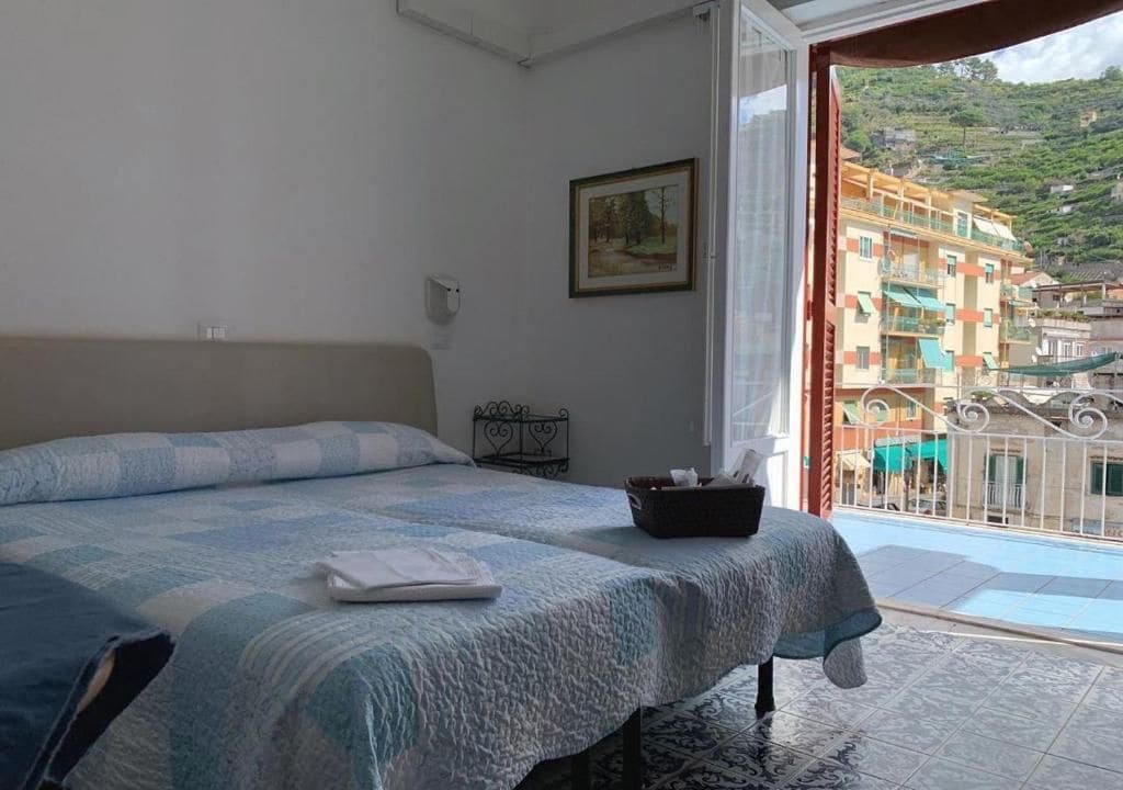 A hotel room with a bed, a small table, a framed picture on the wall, and a balcony with a view of buildings.