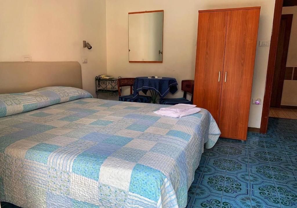 A bedroom with a bed, a wooden wardrobe, a small table with a chair, and a mirror on the wall.