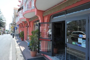 Edifício com entrada por porta de vidro, plantas em vasos e vista clara da rua.
