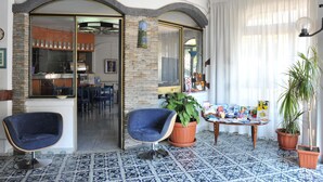 Une pièce avec une chaise bleue, une table avec des magazines, une plante en pot et une vue sur la cuisine.