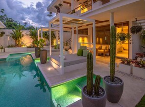 A modern outdoor pool area with a covered seating area, potted plants, and a well-lit interior space.
