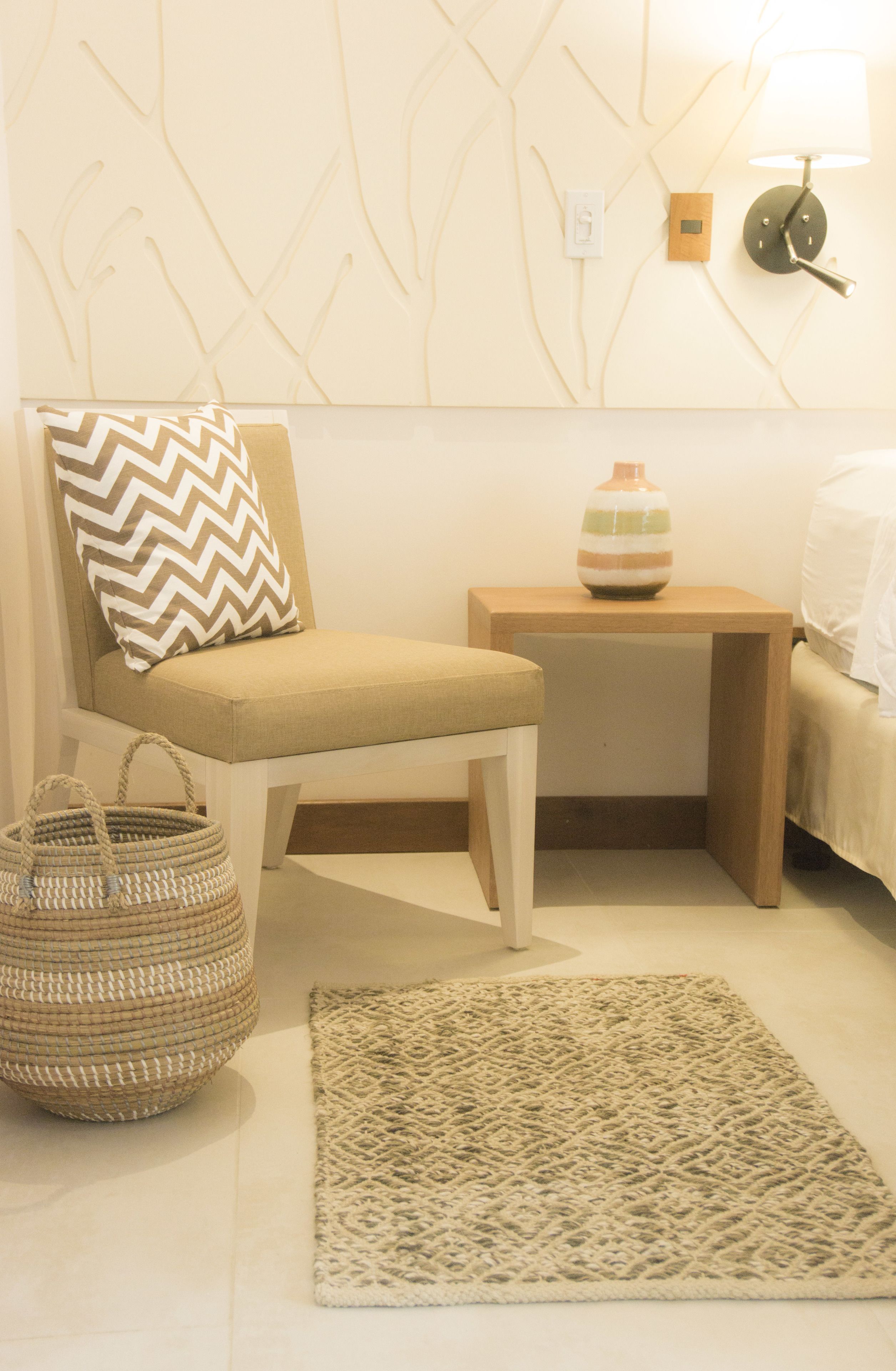 A room with a beige chair, a woven basket, a patterned rug, a bedside table with a lamp, and a wall with a textured design.