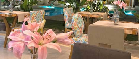 A dining area with tables and chairs, a pool in the background, and potted plants.