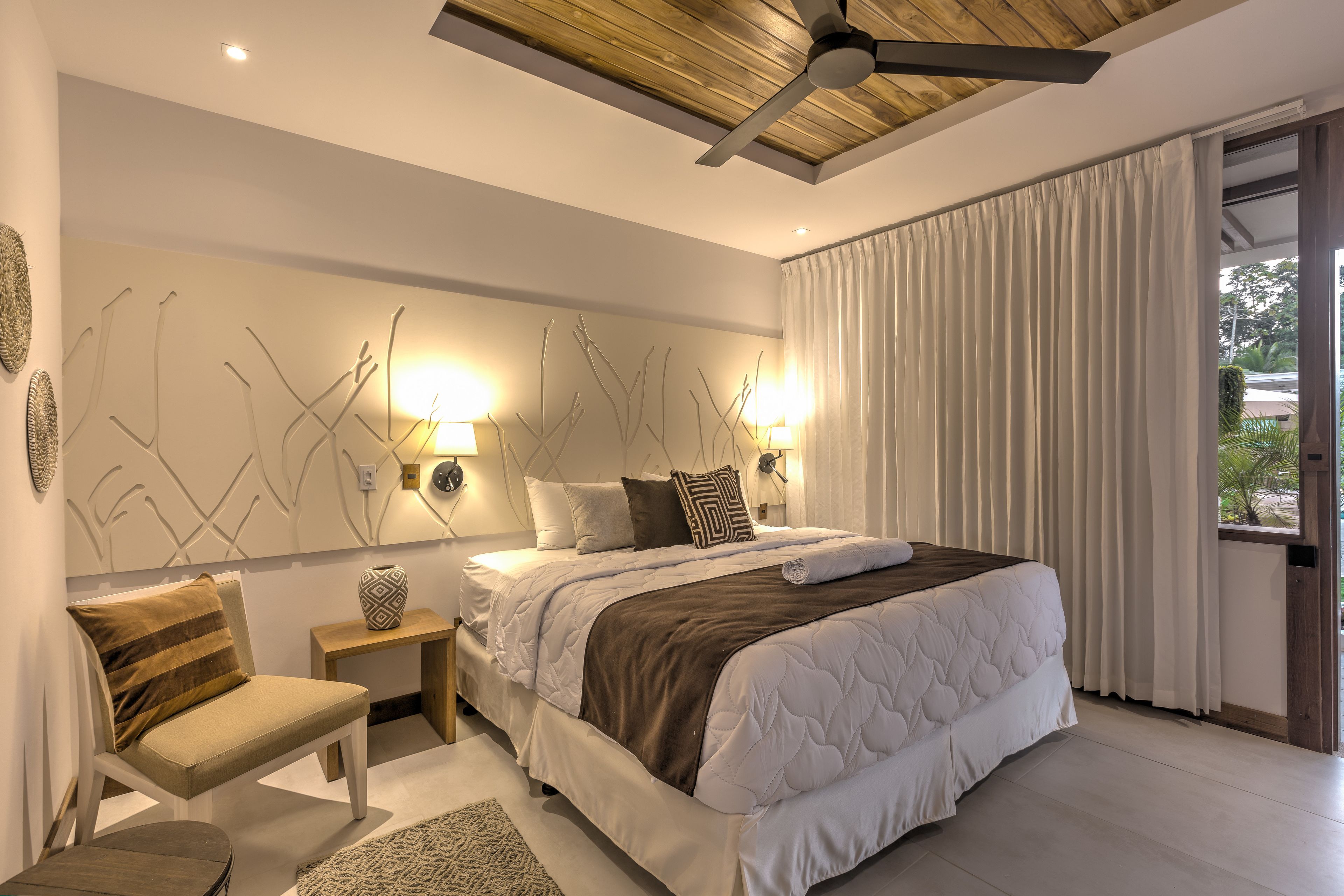A modern bedroom with a large bed, bedside lamps, a ceiling fan, and a view of the outdoors through a window.