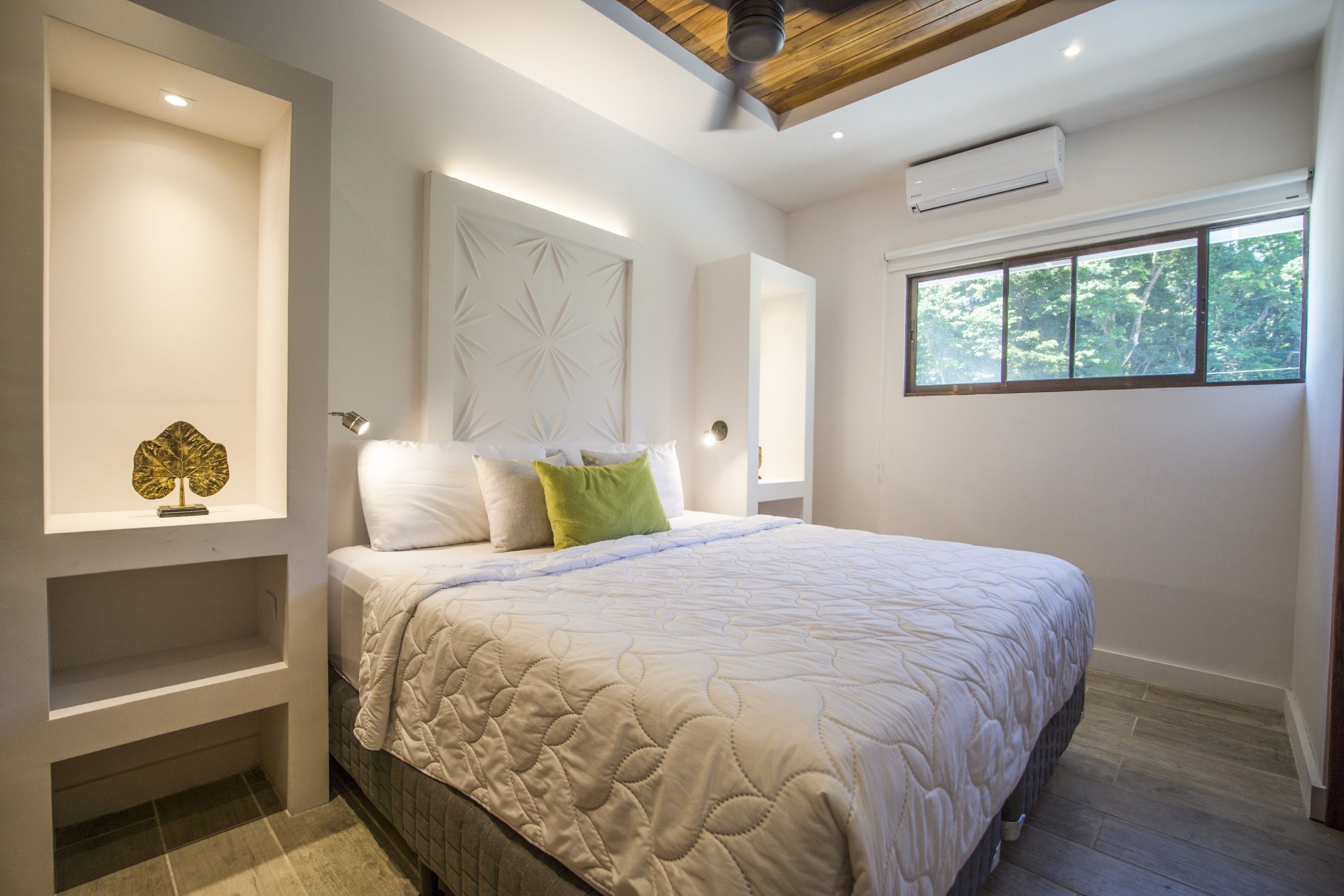 Un dormitorio moderno con una cama grande, estanterías empotradas, ventilador de techo y ventana con vistas a árboles.