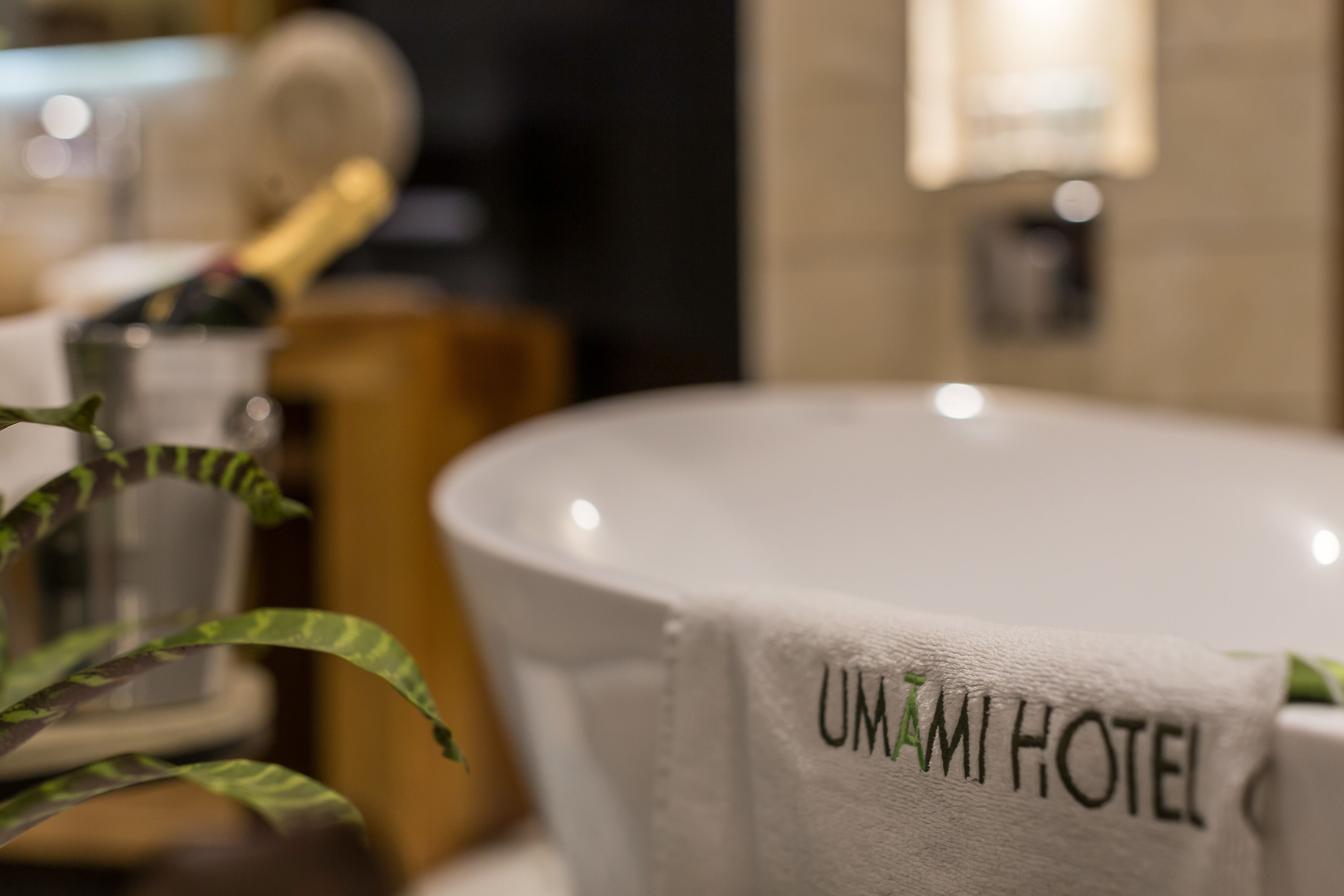 A bathtub with a towel labeled "UMAMI HOTEL" and a plant in the foreground.