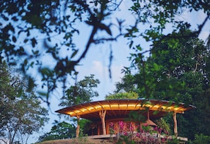 A wooden pavilion with a roof supported by columns, surrounded by lush greenery and trees.