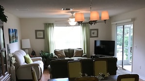Een woonkamer met een bank, een eettafel met stoelen, een televisie en een groot raam met gordijnen.