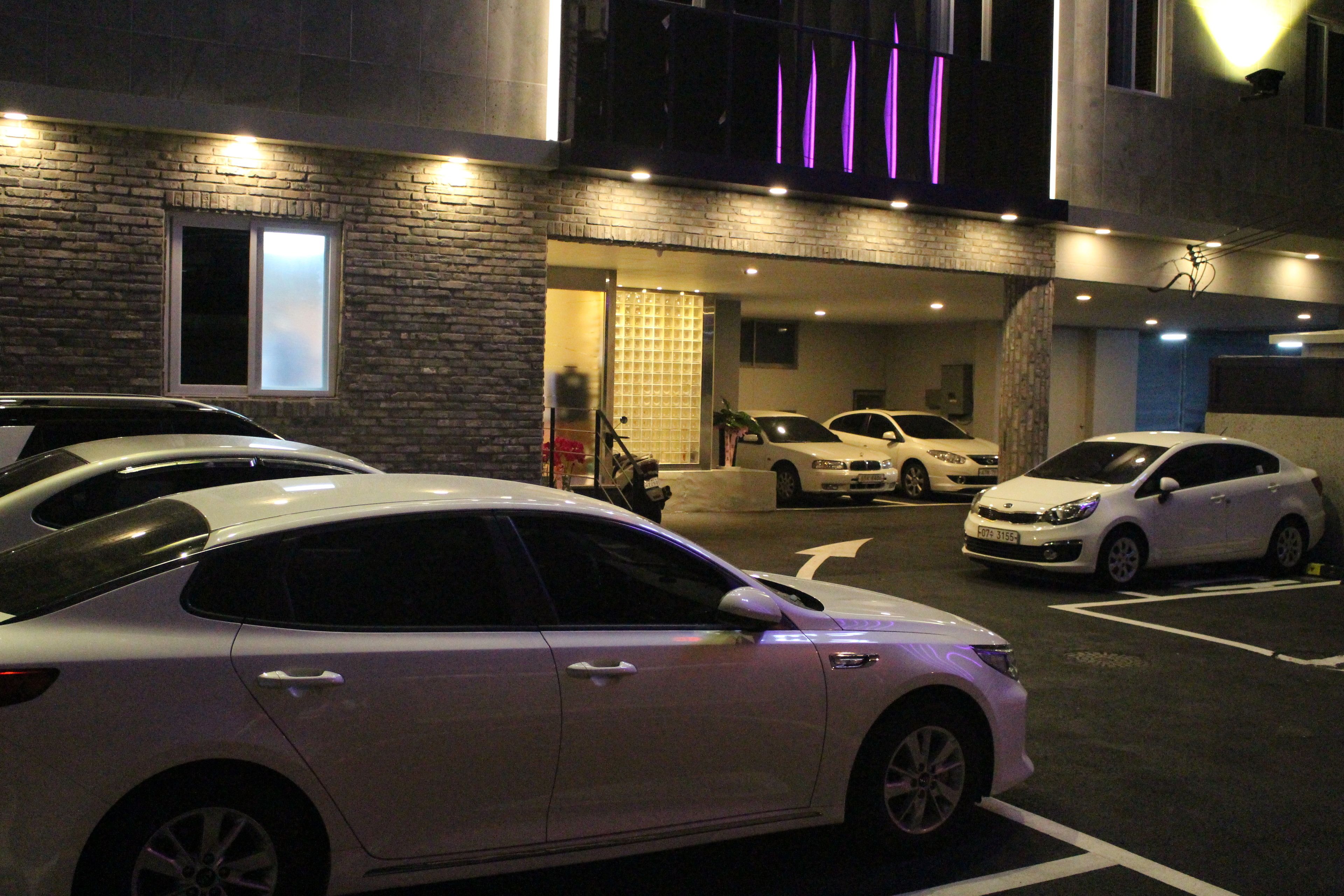 A parking area with several cars, a building entrance with a glass door, and a neon sign above.