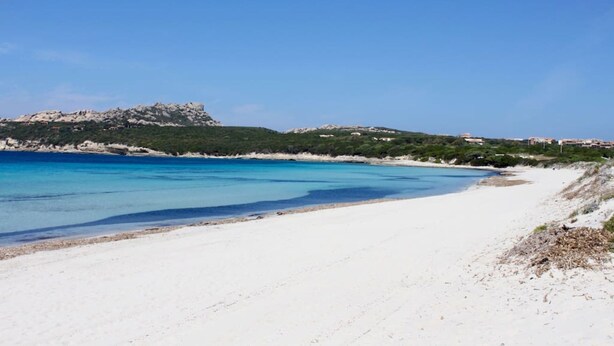 Ein Strand mit weißem Sand, klarem, blauem Wasser und einer felsigen Anhöhe im Hintergrund.