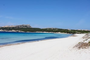 Ein Strand mit weißem Sand, klarem, blauem Wasser und einer felsigen Anhöhe im Hintergrund.