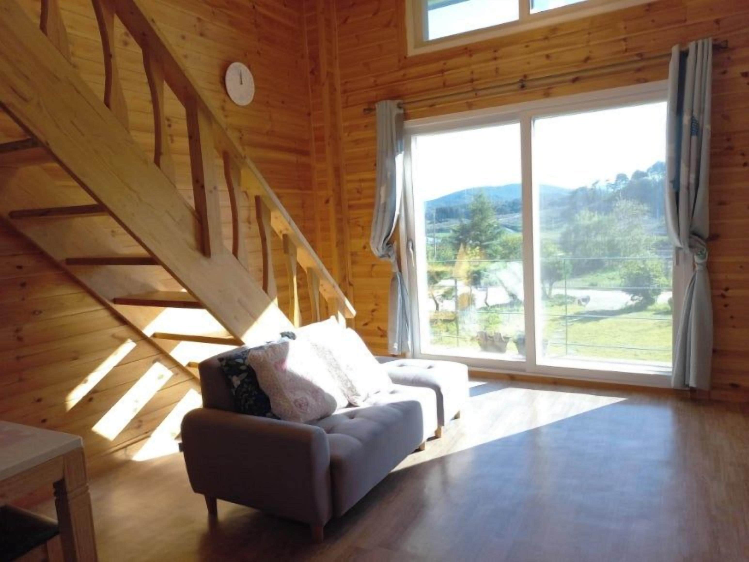 A room with a wooden ceiling, a sofa, a window with curtains, and a view of the outdoors.