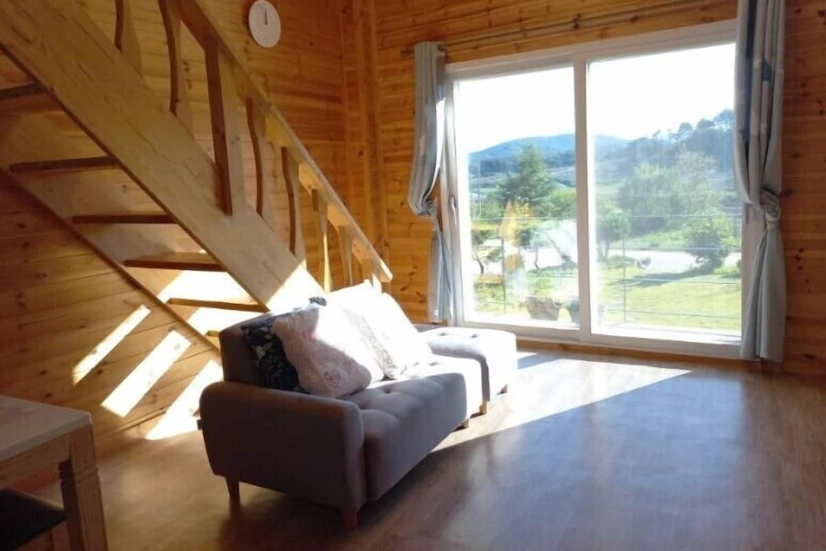 Een houten cabin interieur met een bank, een tafel en een groot raam met uitzicht op een groen landschap.