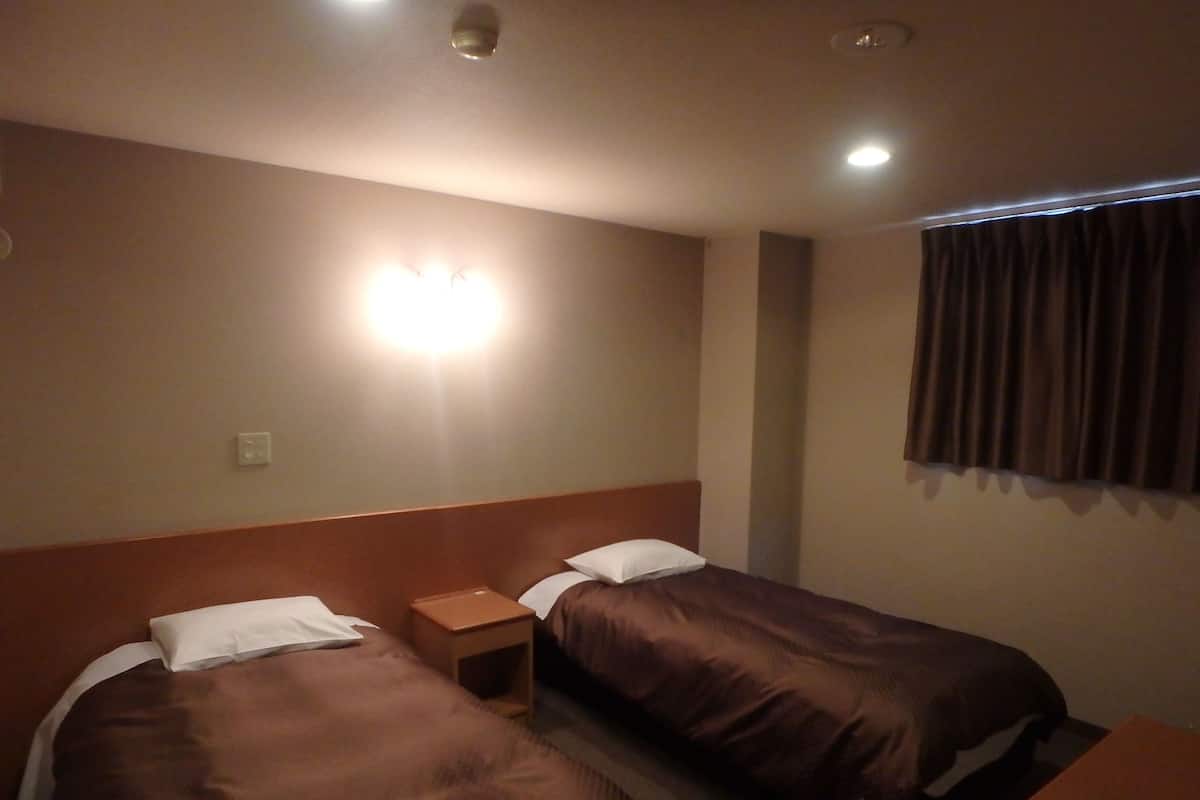 Une chambre d’hôtel avec deux lits, une table de chevet, une lampe fixée au mur et une fenêtre avec des rideaux.