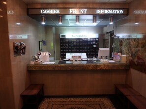 A hotel reception desk with a 'CASHIER' sign, a 'FRONT INFORMATION' sign, and a computer monitor. There are brochures and a small clock on the desk. - Hotel SUNTARGAS UENO (Tokyo)