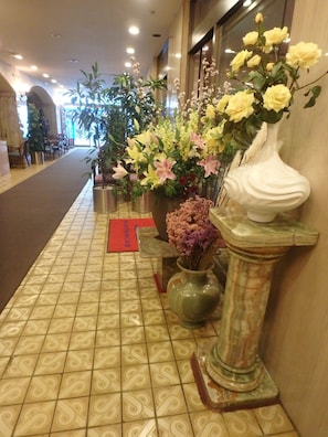 A hallway with a patterned tile floor, a reception desk, and various potted plants including flowers and a large vase. - Hotel SUNTARGAS UENO (Tokyo)