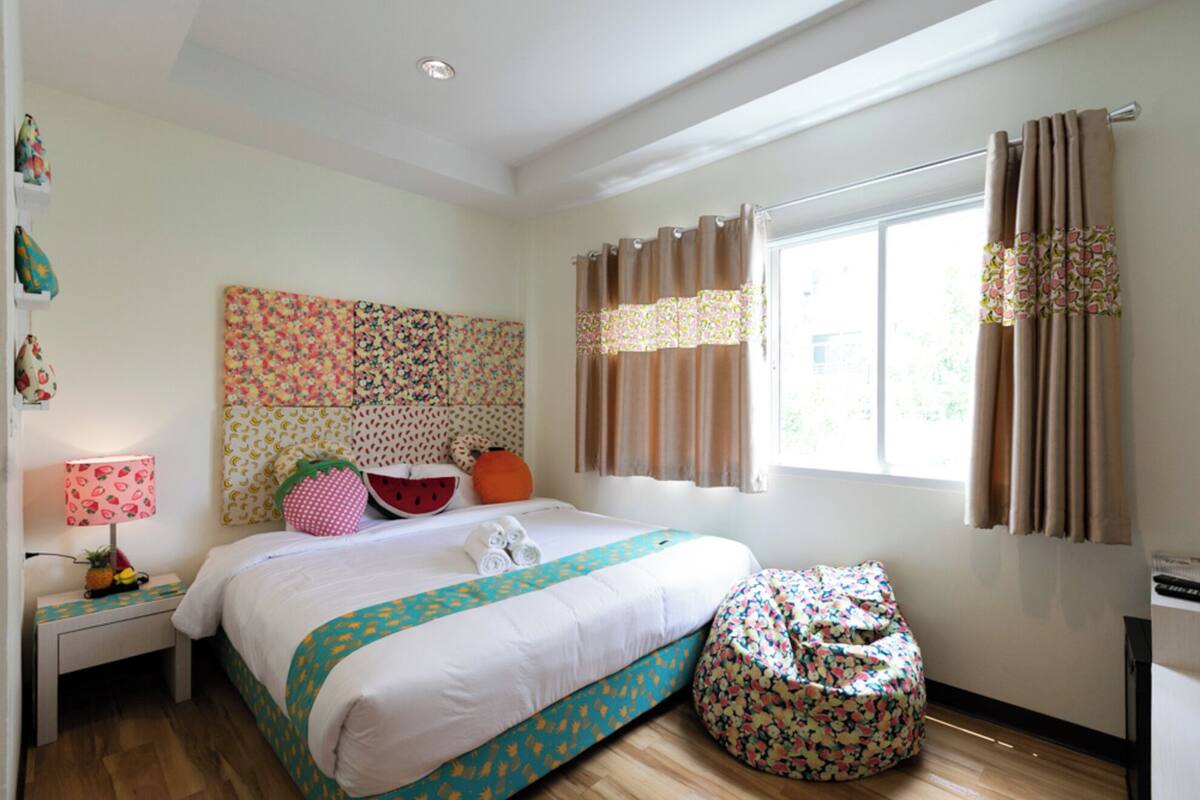 Une chambre à coucher avec un lit, une table de chevet avec une lampe, une fenêtre avec des rideaux et un fauteuil en forme de poire.