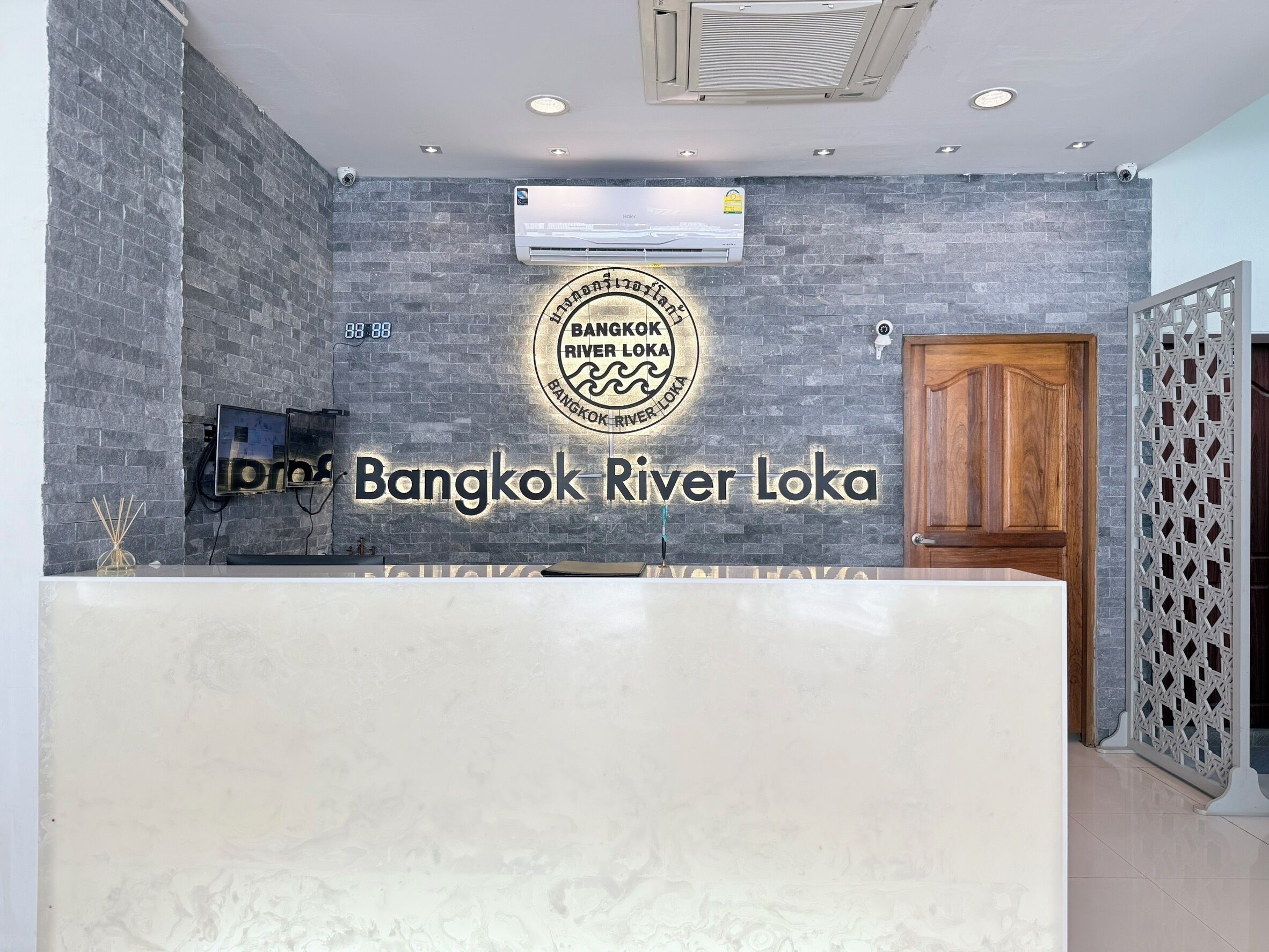 A reception desk with a logo for Bangkok River Loka, a modern interior with grey brick walls, a wooden door, and a security camera.