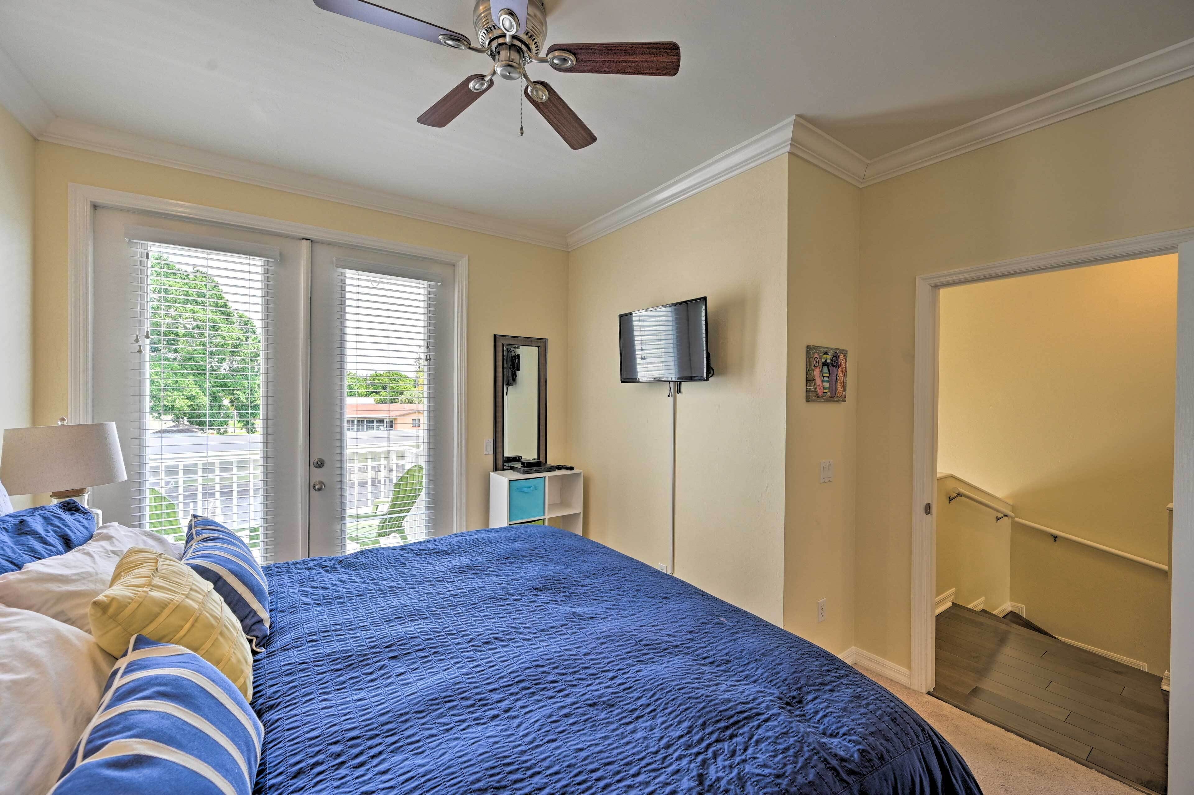 A bedroom with a bed, a ceiling fan, a TV, and a door leading to a balcony.