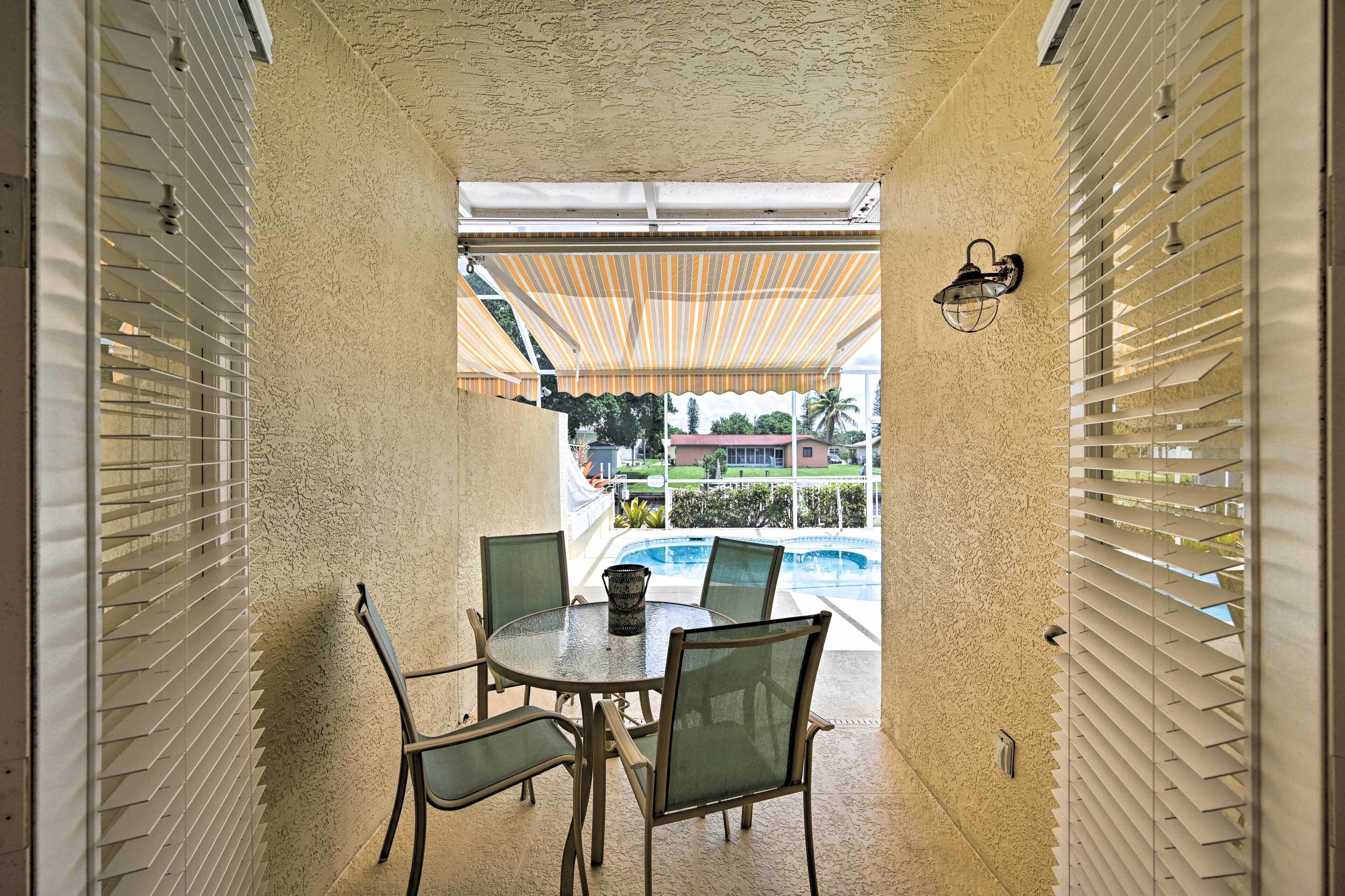 A covered outdoor dining area with a table and chairs, a view of a swimming pool, and a building in the background.