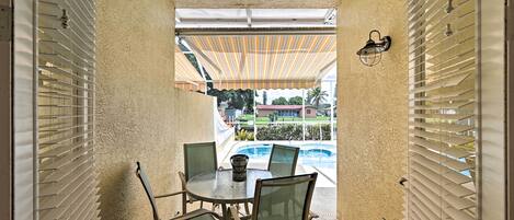 A covered outdoor dining area with a table and chairs, a view of a swimming pool, and a building in the background.