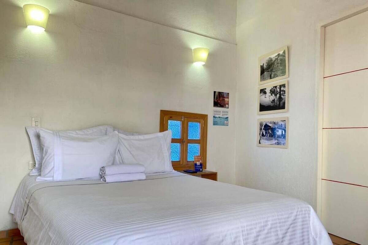 Una cama bien hecha con sábanas blancas, una mesita de noche de madera con una lámpara, y una pared con cuadros enmarcados.