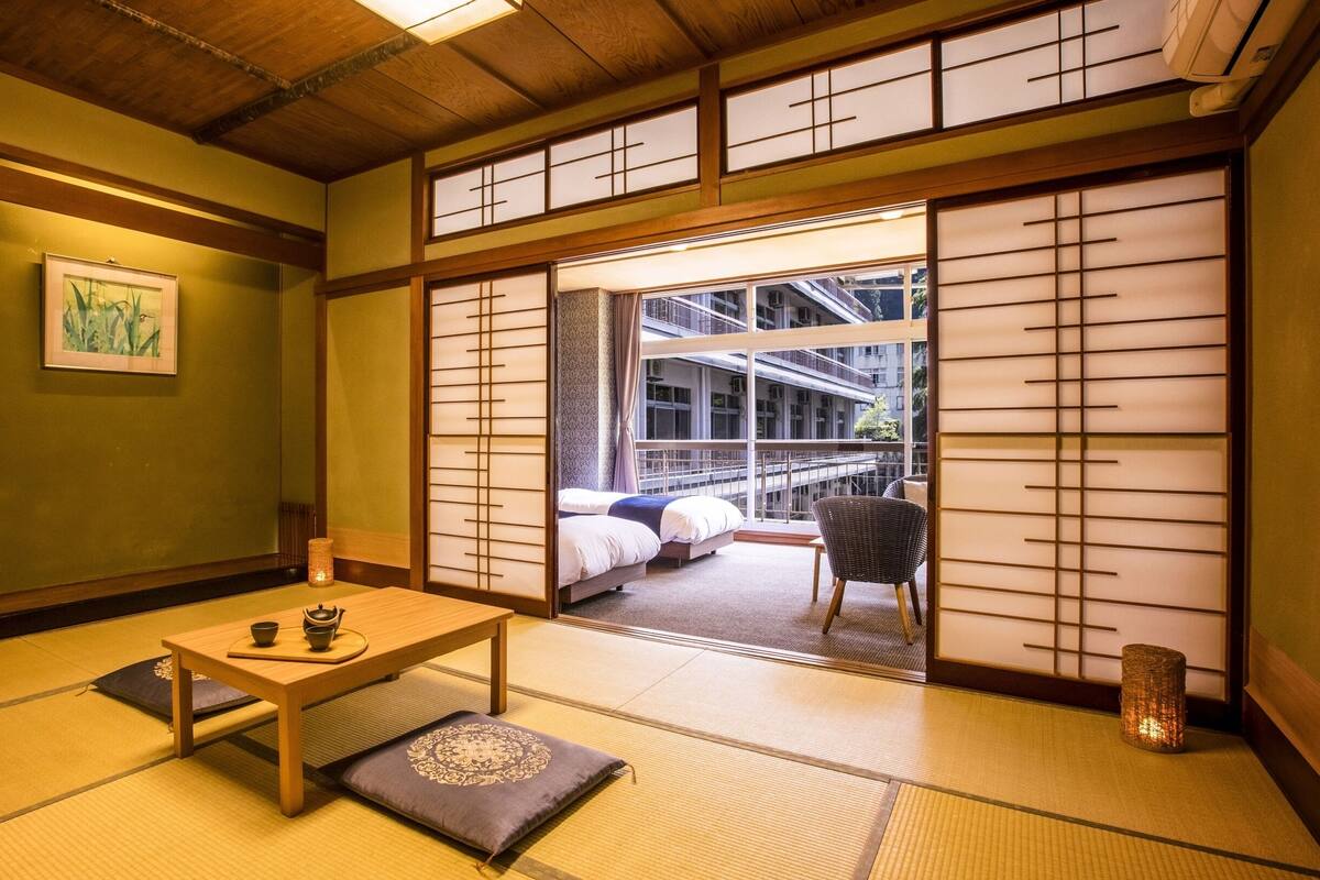 Une pièce de style japonais traditionnel, avec un sol recouvert de tatamis, une petite table sur laquelle est disposé un service à thé, et une vue sur un bâtiment à travers des panneaux coulissants en papier.