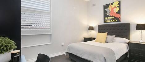 A modern bedroom with a bed, bedside tables, a window with blinds, a potted plant, and a poster on the wall.