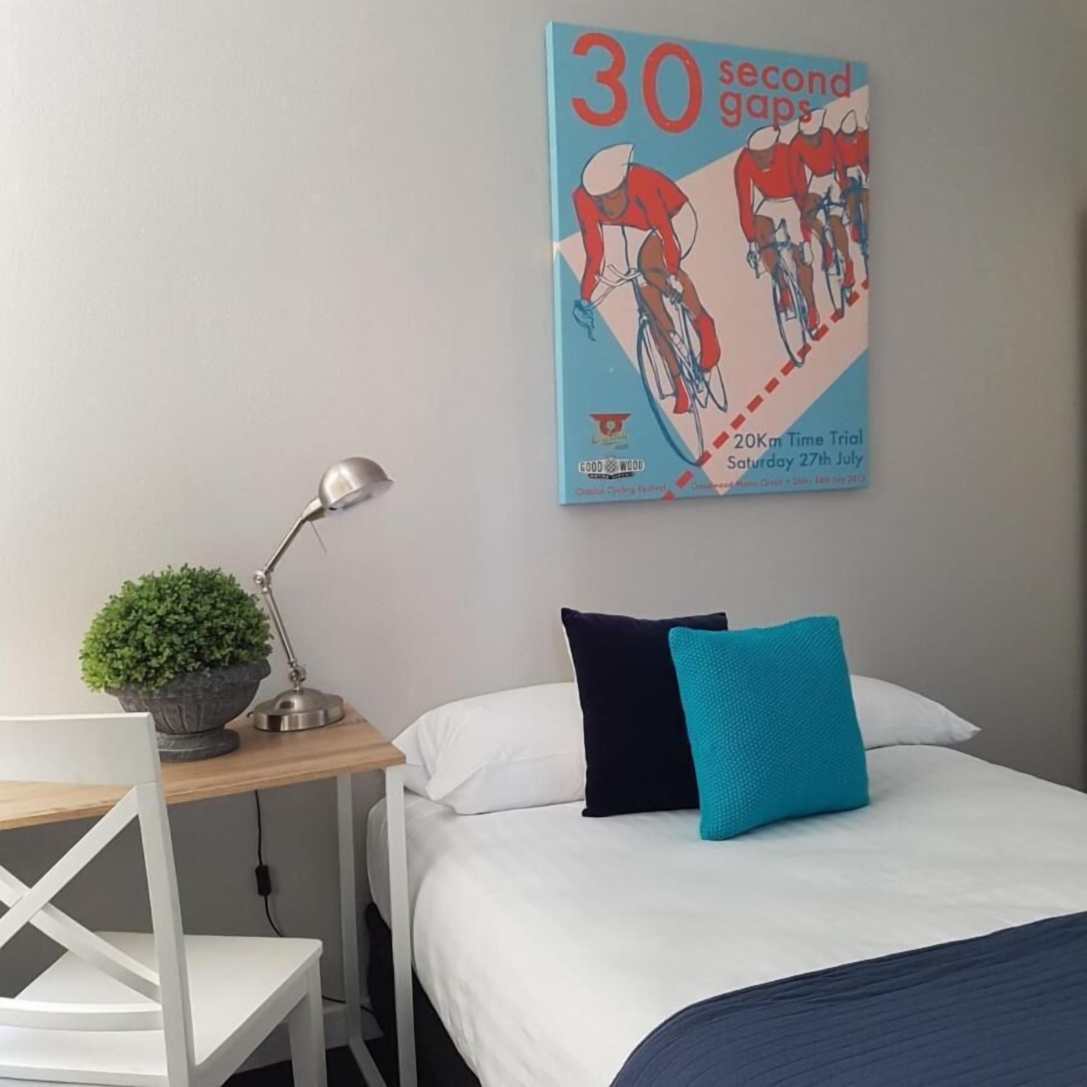 A hotel room with a bed, a desk with a lamp, and a poster on the wall advertising a cycling event.