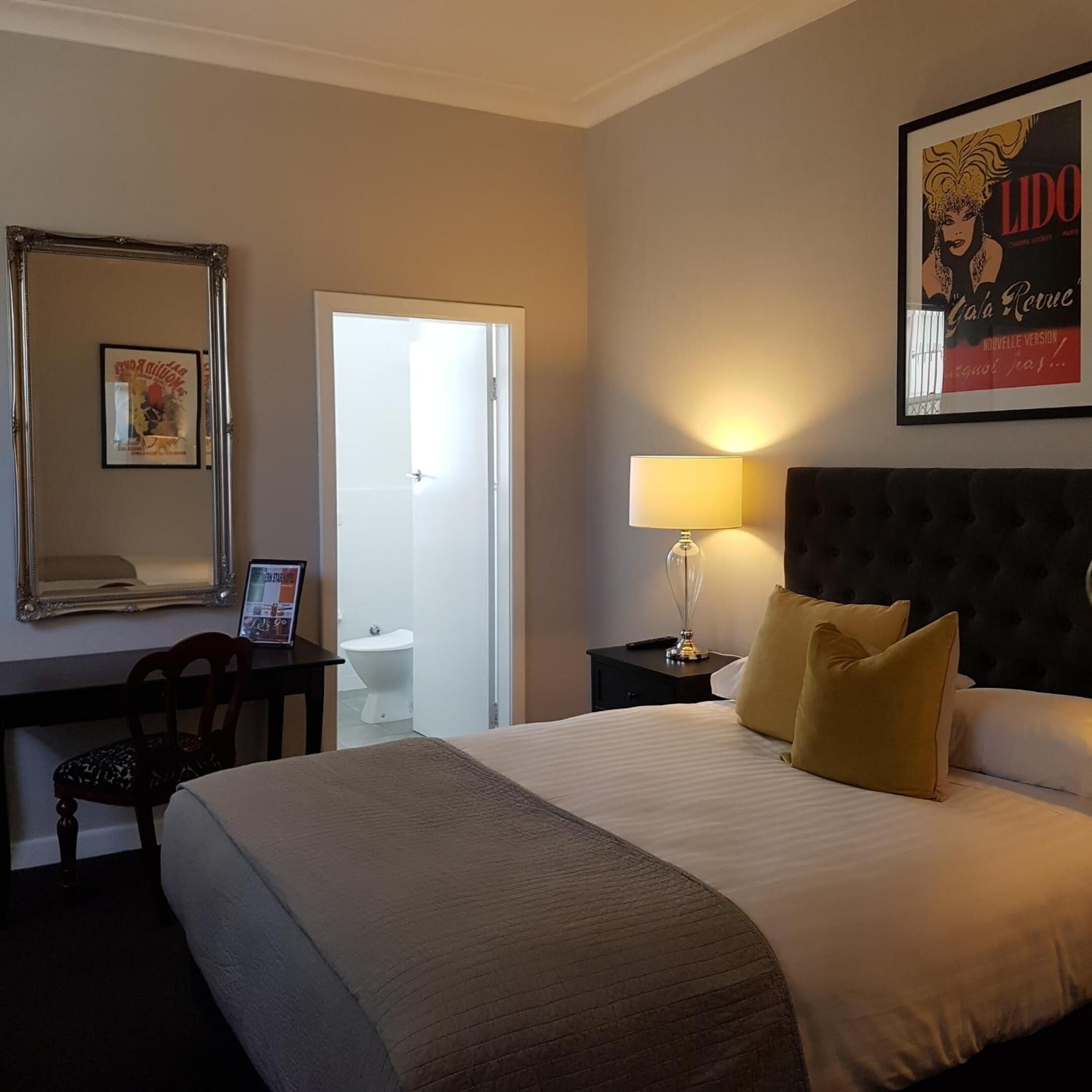 Ein Hotelzimmer mit einem großen Bett, einem Schreibtisch mit Stuhl, einem Nachttisch mit Lampe, einem Spiegel und einem gerahmten Poster an der Wand.