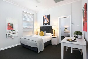 A modern hotel room with a large bed, bedside tables with lamps, a small desk, and artwork on the walls.