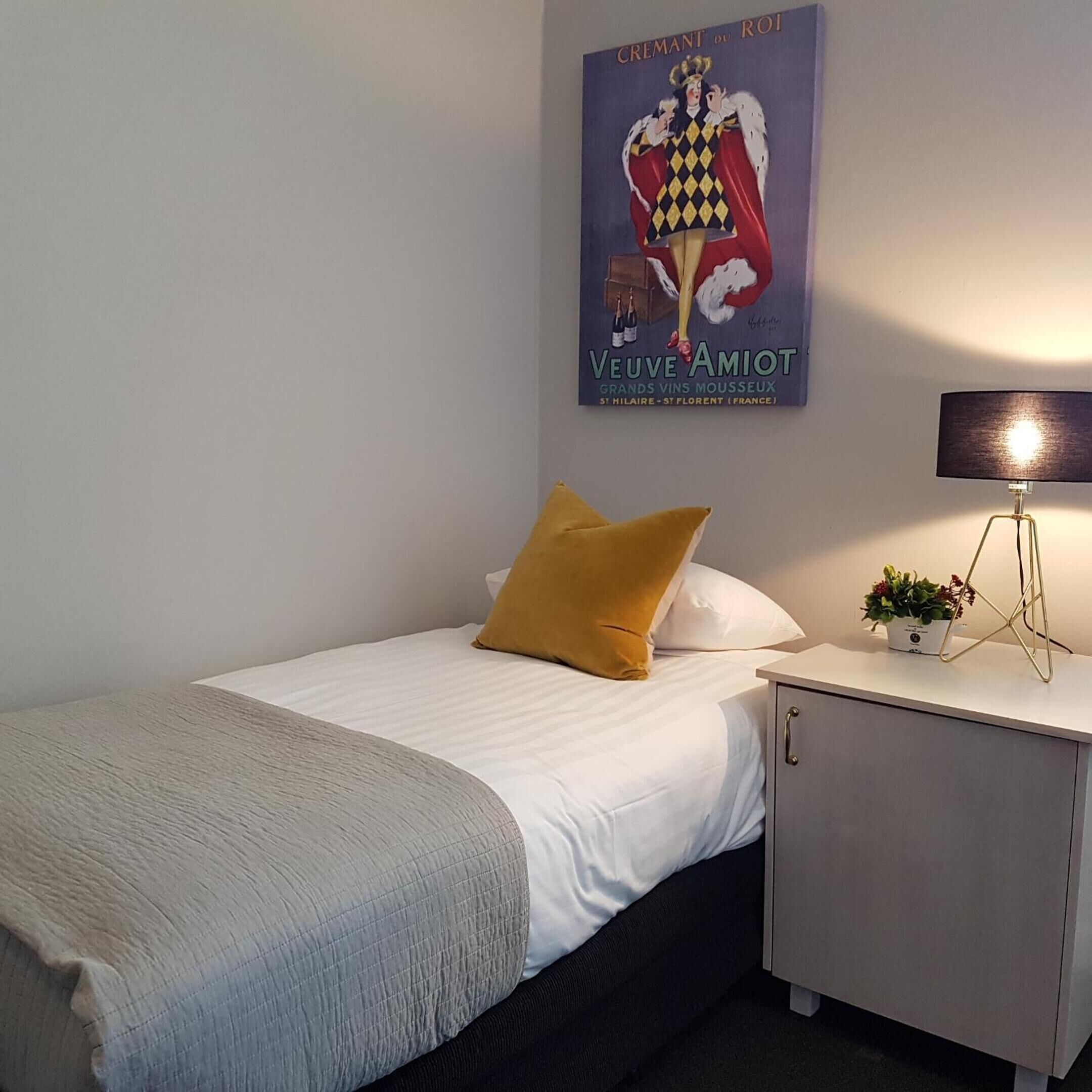 A neatly made bed with a white comforter and a mustard-colored pillow, a bedside table with a lamp and a small plant, and a poster on the wall advertising Veuve Amiot wine.