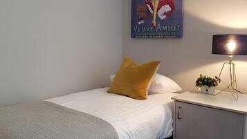Una cama bien hecha con edredón blanco y almohada color mostaza, una mesita de noche con lámpara y una pequeña planta, y un cartel en la pared que anuncia vino Veuve Amiot.